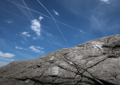 Das Foto zeigt einen großen Felsbrocken mit Riefen, deren Muster sich in den Kondensstreifen am Himmel fortsetzt (Weg zur Kreuzspitze im Ratschingstal, Südtirol) - fotografiert im Juli 2022